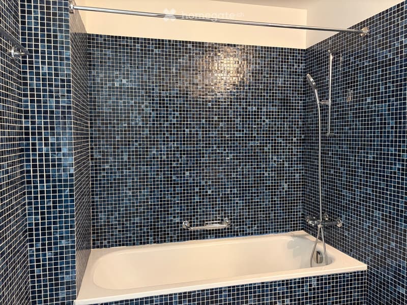 Bathtub with modern dark blue tiles, showerhead, and handrail.