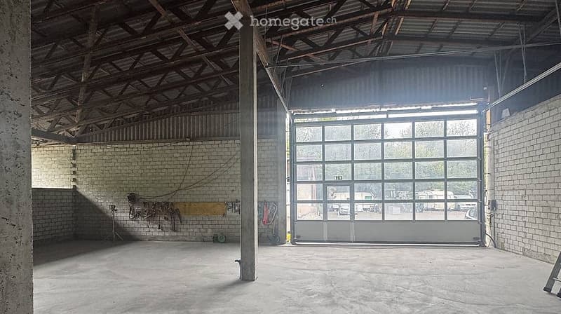 Empty garage with exposed wooden beams, brick walls, and a large roll-up door. No furniture or equipment present.