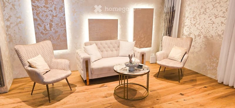 Modern living room with a white tufted sofa, two armchairs, a round coffee table, and decorative items on the table. The room features wooden flooring and large windows with curtains.