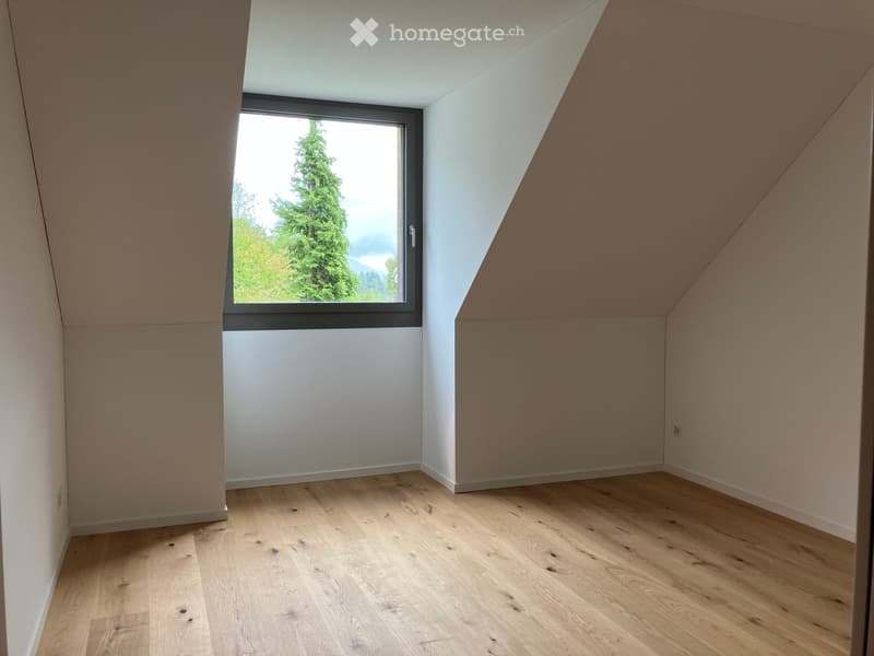 Empty attic room with wooden flooring, white walls, and a large window offering a view of greenery outside.