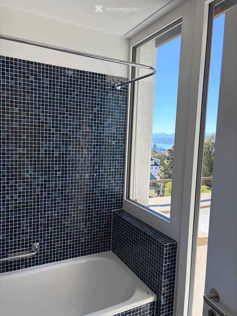 Modern bathroom with a large window offering a scenic view, featuring a bathtub and a shower handle.