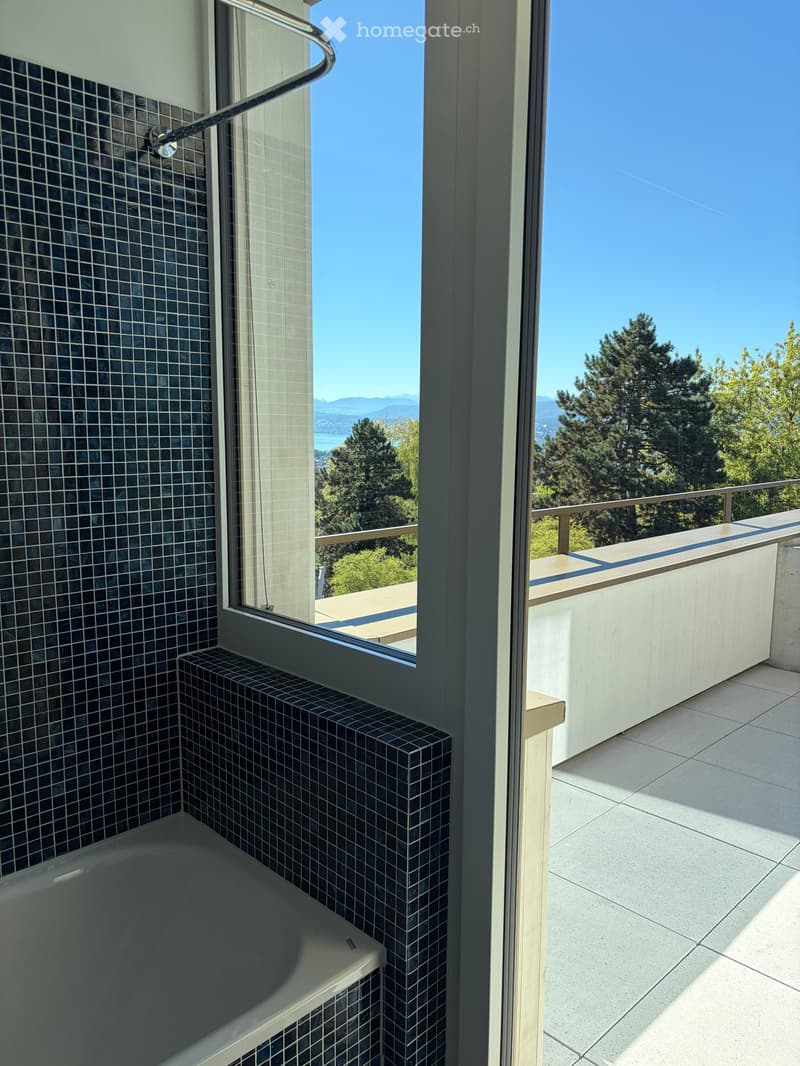 Bathroom with a bathtub, dark tiled walls, and a large window opening to a balcony with a scenic view of trees and mountains.