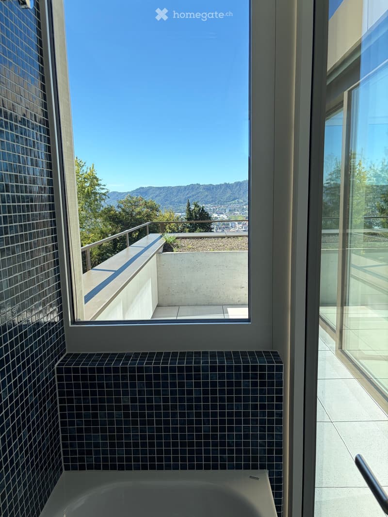 Modern bathroom with a bathtub, black tiled walls, and a large window offering a scenic view of the city and mountains.