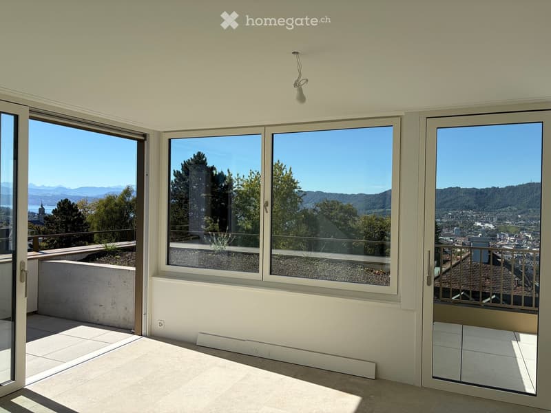 Empty room with large windows and sliding doors leading to a balcony, offering scenic views of trees and cityscape.