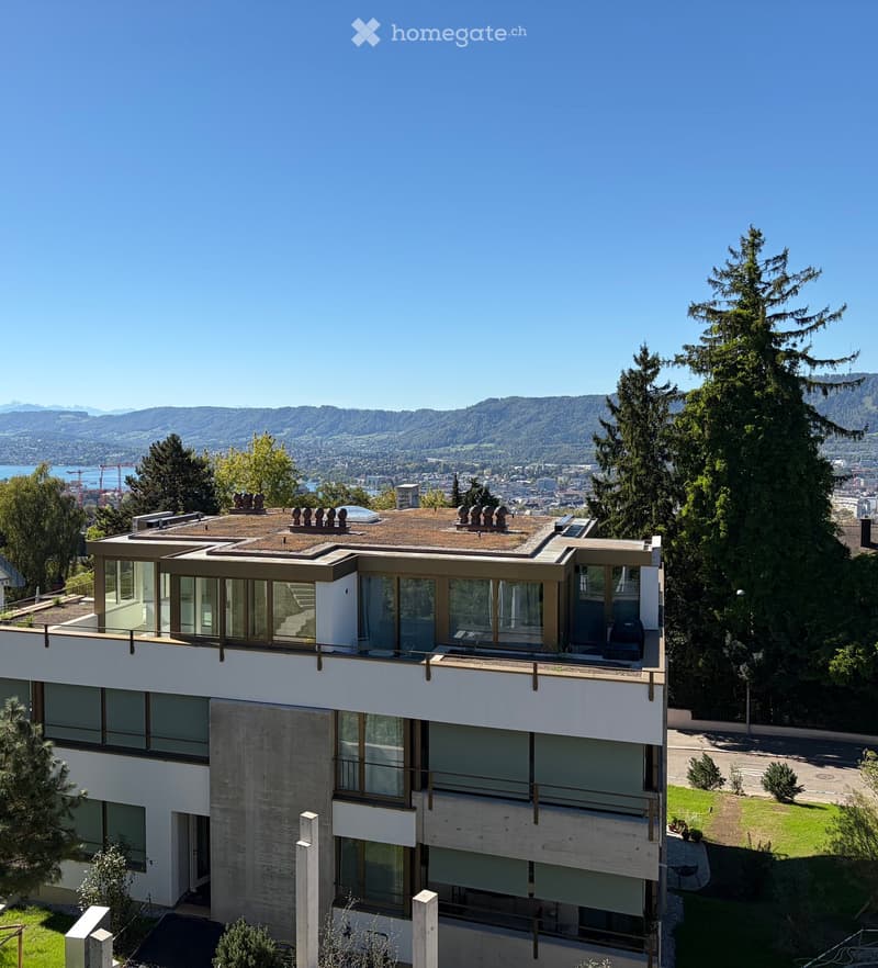 Modern multi-story building with large windows and a flat roof, surrounded by trees and offering a scenic view of a city and mountains.