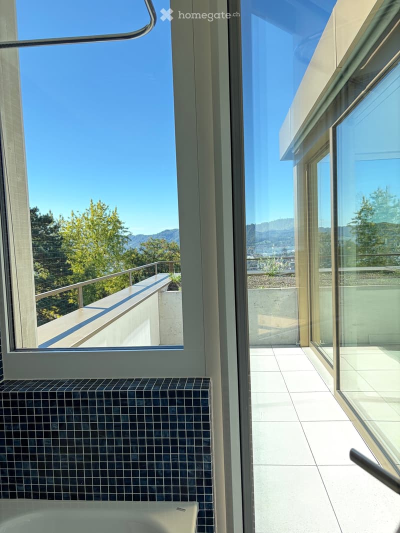 Modern bathroom with a bathtub, large windows offering a scenic view of trees and mountains, and a tiled floor.