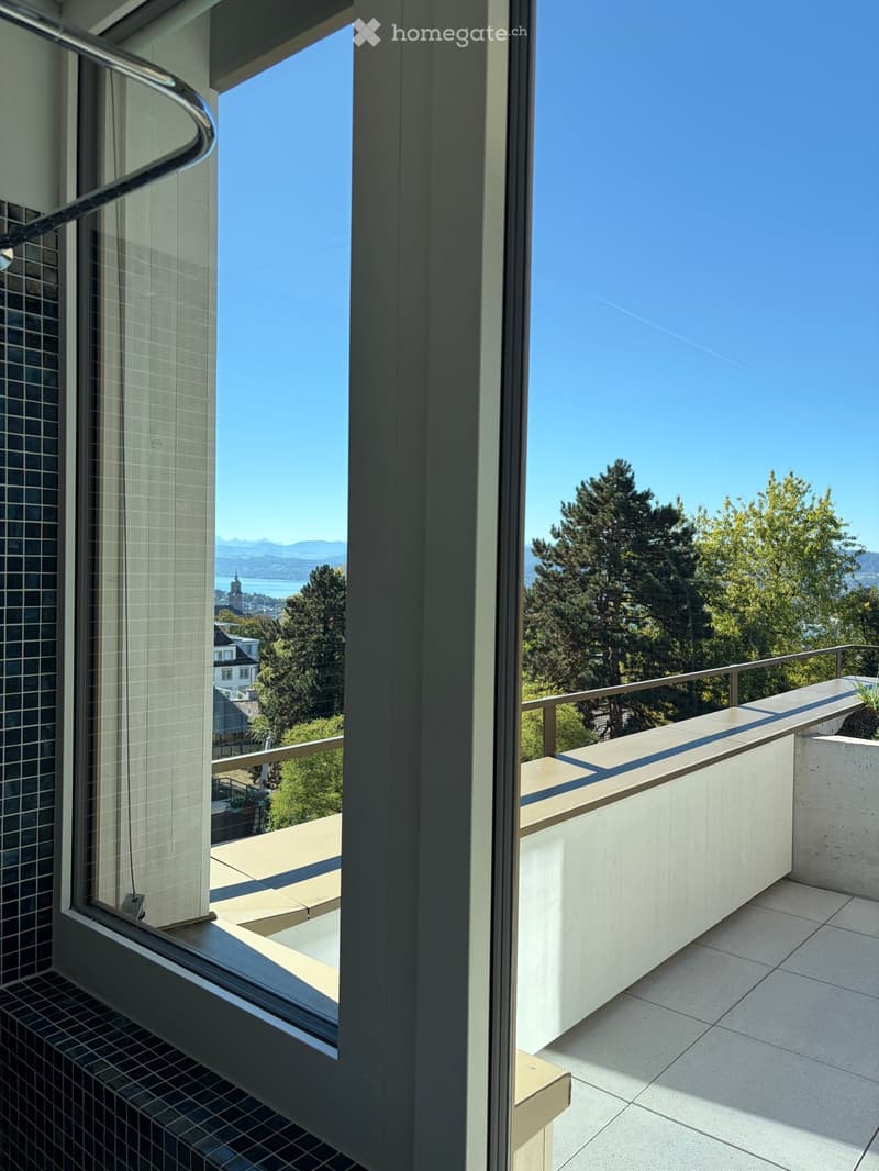 Modern balcony with concrete seating area, metal railing, and scenic view of trees and distant water.