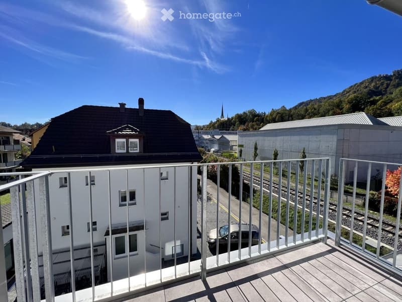 Balcony with metal railing overlooking a street, train tracks, and surrounding buildings under a clear blue sky.