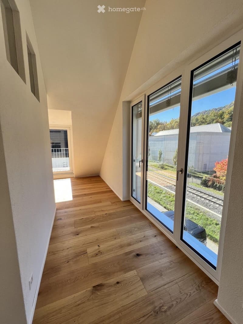 An empty room with wooden flooring and large windows offering a view of the outdoors, including a grassy area and a parked car.