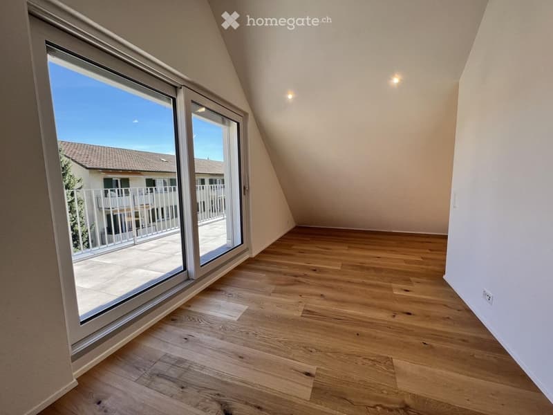 Empty attic room with wooden flooring, large window offering a view of a balcony and neighboring house.