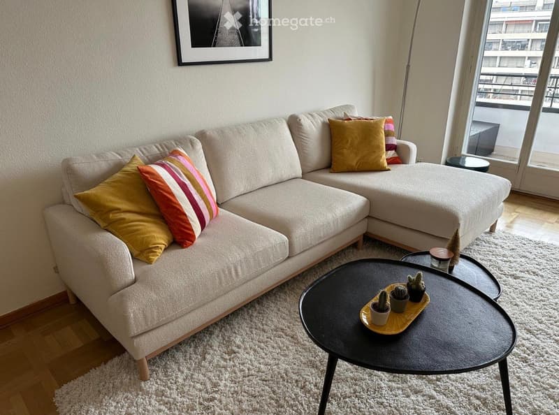 Modern living room with a beige sectional sofa, colorful throw pillows, a black coffee table with small potted plants, a floor lamp, and a view of a cityscape through large windows.