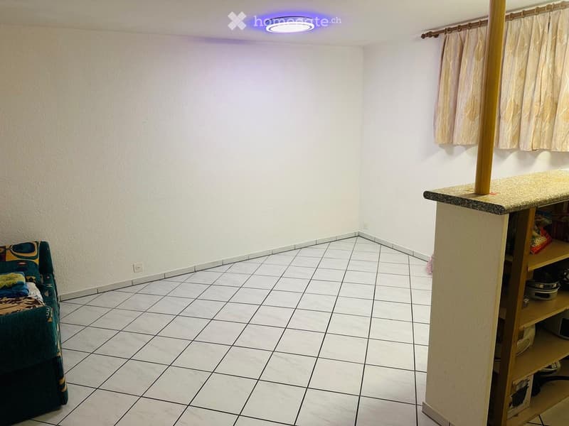 A small, furnished room with a tiled floor, white walls, a sofa with colorful cushions, a small table with a countertop, and shelves holding various items.