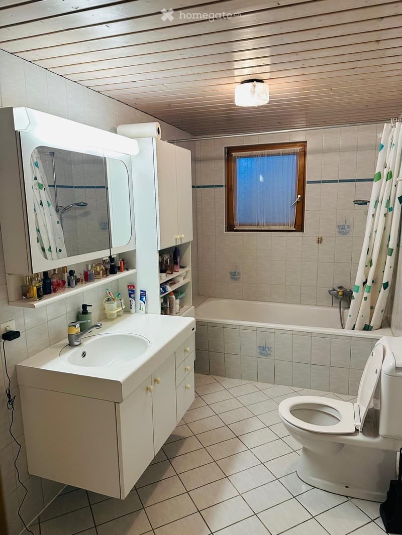 Bathroom with a white sink, mirror, cabinet, toilet, bathtub, and shower curtain. The room features tiled walls and floor, a window, and a light fixture on the ceiling.