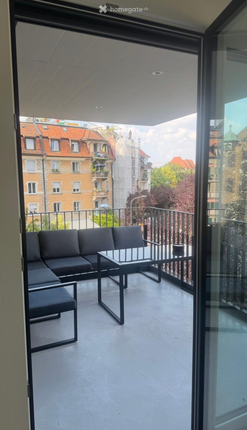 Modern balcony with a grey sectional sofa, a glass coffee table, and a small black bowl on the table. The balcony has a metal railing and overlooks a cityscape with historic buildings.