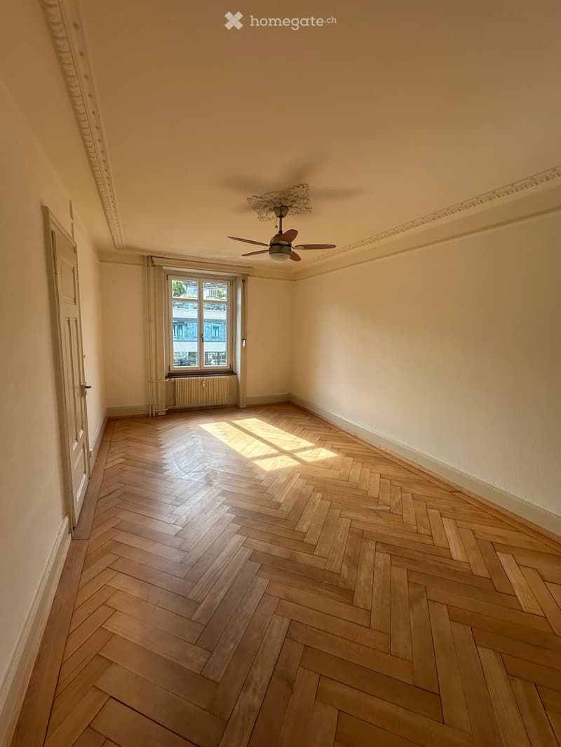 Empty room with herringbone wooden flooring, white walls, a ceiling fan, and a window with a radiator below.