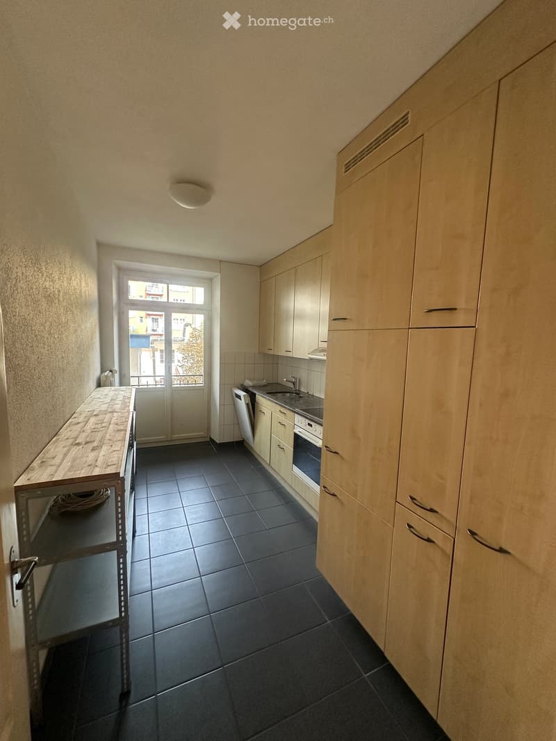 Modern kitchen with light wood cabinets, dark tiled floor, sink, oven, dishwasher, and a wooden table.