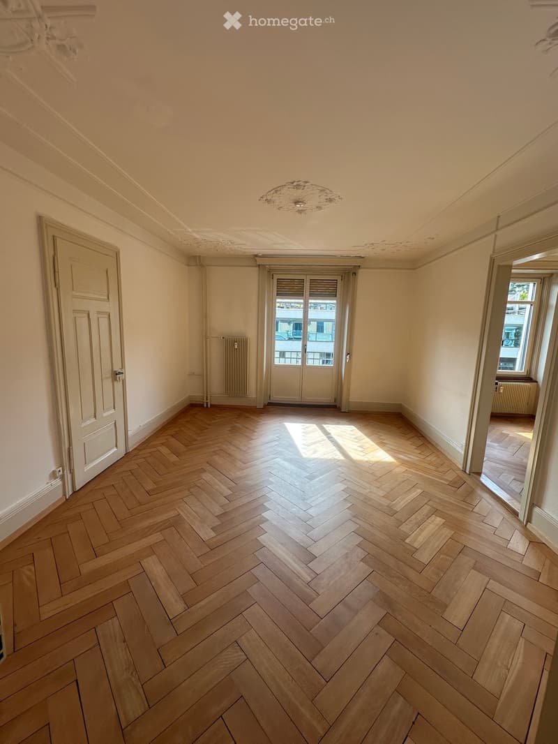 Empty room with herringbone wooden flooring, white walls, and a decorative ceiling light fixture. Features a door, a windowed entryway, and radiators on both sides.