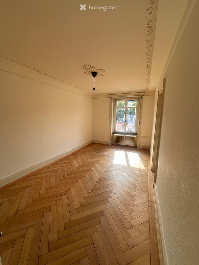 Empty room with herringbone wooden flooring, white walls, and a single window with radiator below. Natural light streams in from the window.