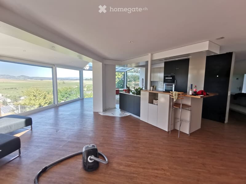 Modern kitchen and living area with large windows, wooden flooring, kitchen island, stools, and a sofa.