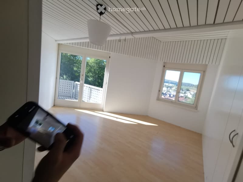 Empty room with wooden flooring, white walls, large windows, and a view of greenery and cityscape.