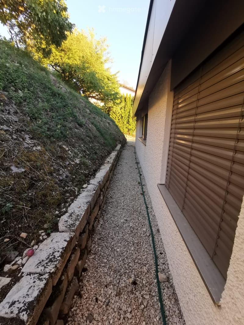A narrow, elevated pathway between a house and a sloped garden area, covered in gravel and bordered by stone walls.