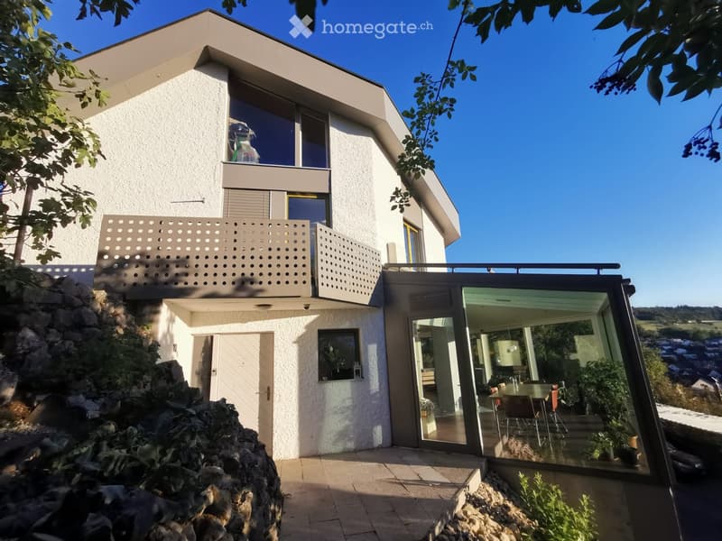 Modern two-story house with white exterior, large windows, balcony, and glass-enclosed patio area with dining table and chairs.