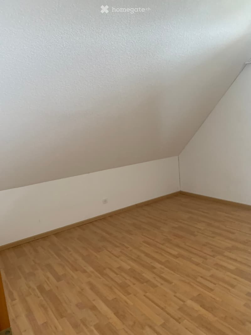 Empty room with wooden flooring and white walls, featuring a slanted ceiling and a visible electrical outlet.