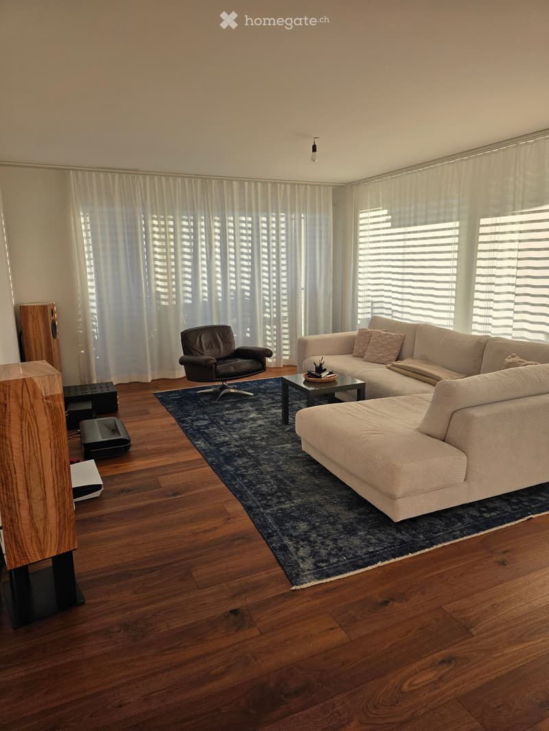 Modern living room with wooden flooring, large windows with white curtains, a sectional sofa, armchair, coffee table, speaker system, and a blue rug.