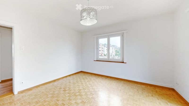 Empty room with wooden flooring, white walls, and a single window with a view of buildings outside. A modern pendant light hangs from the ceiling.
