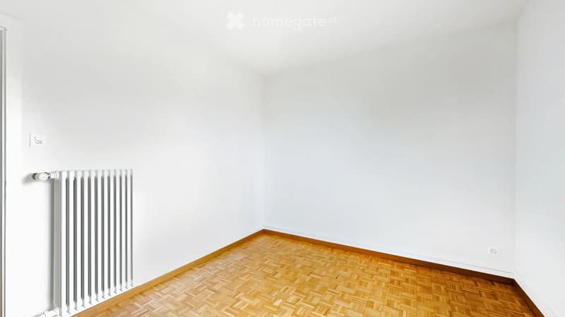 Empty room with white walls, wooden herringbone floor, radiator, and electrical outlet.