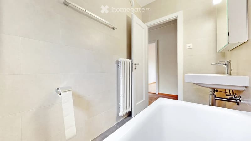 Modern bathroom with a white bathtub, sink, radiator, and mirror. The walls are tiled in light beige, and the floor is tiled in dark grey.