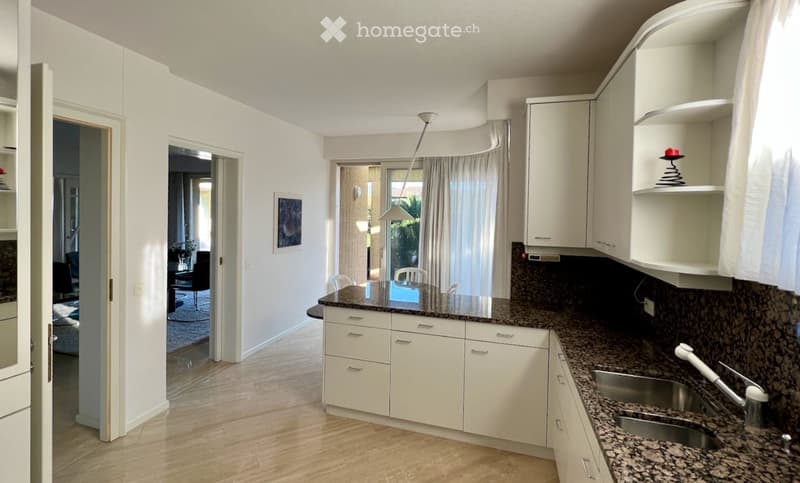 Modern kitchen with white cabinets, granite countertops, and a stainless steel sink. Large window with a view, pendant light fixture, and an open doorway leading to another room.