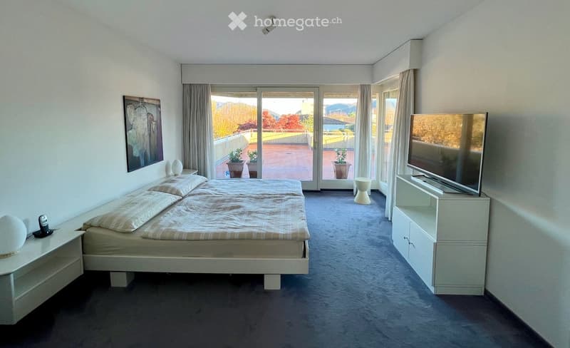 Modern bedroom with a large bed, nightstands, lamps, TV stand, and a view of the outdoor area through large windows.