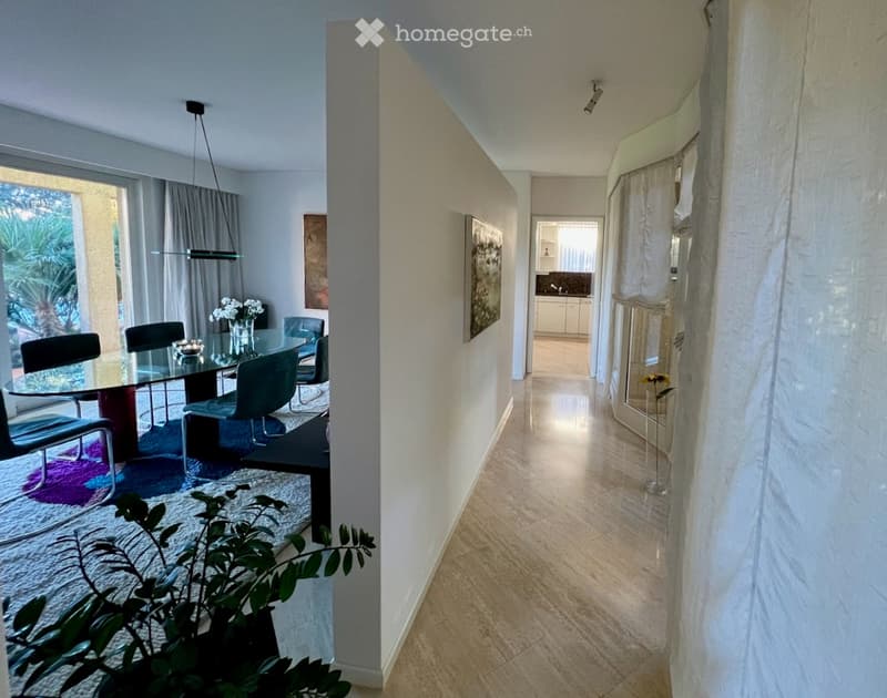 Modern dining area with a glass table, chairs, and a vase of flowers, connected to a hallway leading to a kitchen area with cabinets and a countertop.