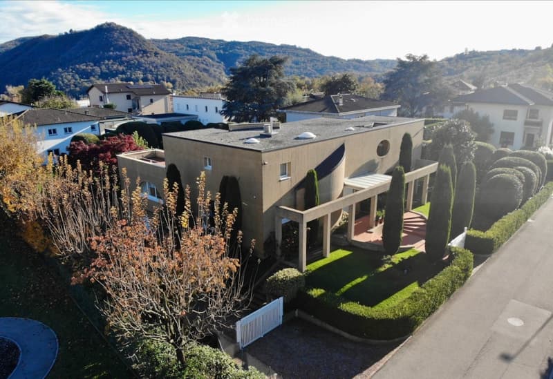 Modern two-story house with a flat roof, large windows, and a spacious garden with neatly trimmed hedges and trees. The house is surrounded by other residential buildings and features a scenic mountain view in the background.