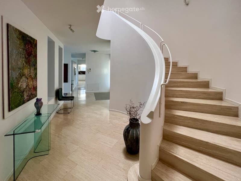 Spacious hallway with a curved staircase, glass console table, two chairs, and decorative vases.