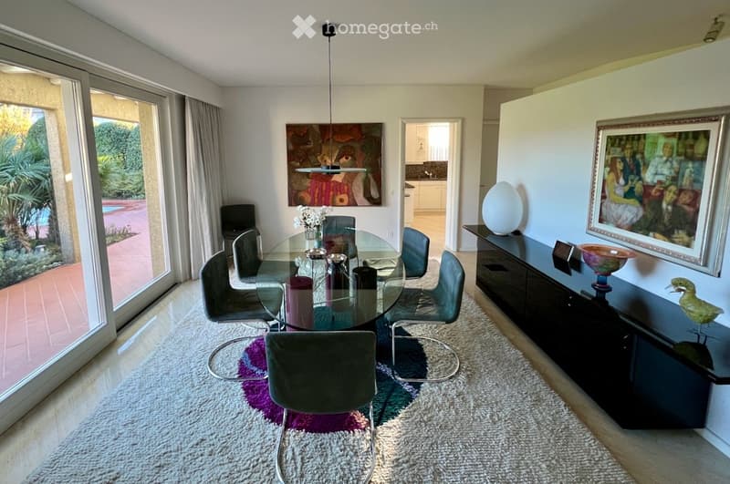 Dining room with a glass dining table, six chairs, a dark wooden cabinet, a white vase, a colorful picture frame, a decorative bowl, and a decorative bird on the cabinet.