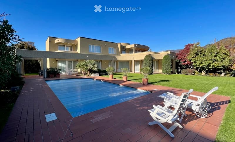 A modern two-story building with a swimming pool in the foreground, surrounded by well-maintained lawn, potted plants, and white lounge chairs.
