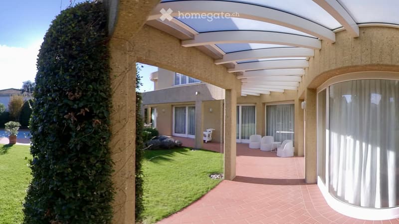 A covered outdoor patio area with white furniture, surrounded by a well-maintained lawn and a hedge. The patio has a brick floor and is connected to a house with large windows and a modern design.