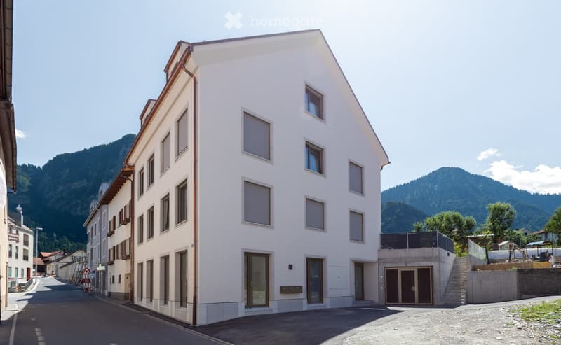 Modern white building with large windows and a raised ground floor entrance, situated in a scenic town with mountains in the background.