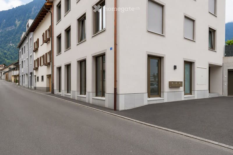 Modern multi-story building with white facade, large windows, and balconies, situated in a quiet street with a scenic mountain backdrop.