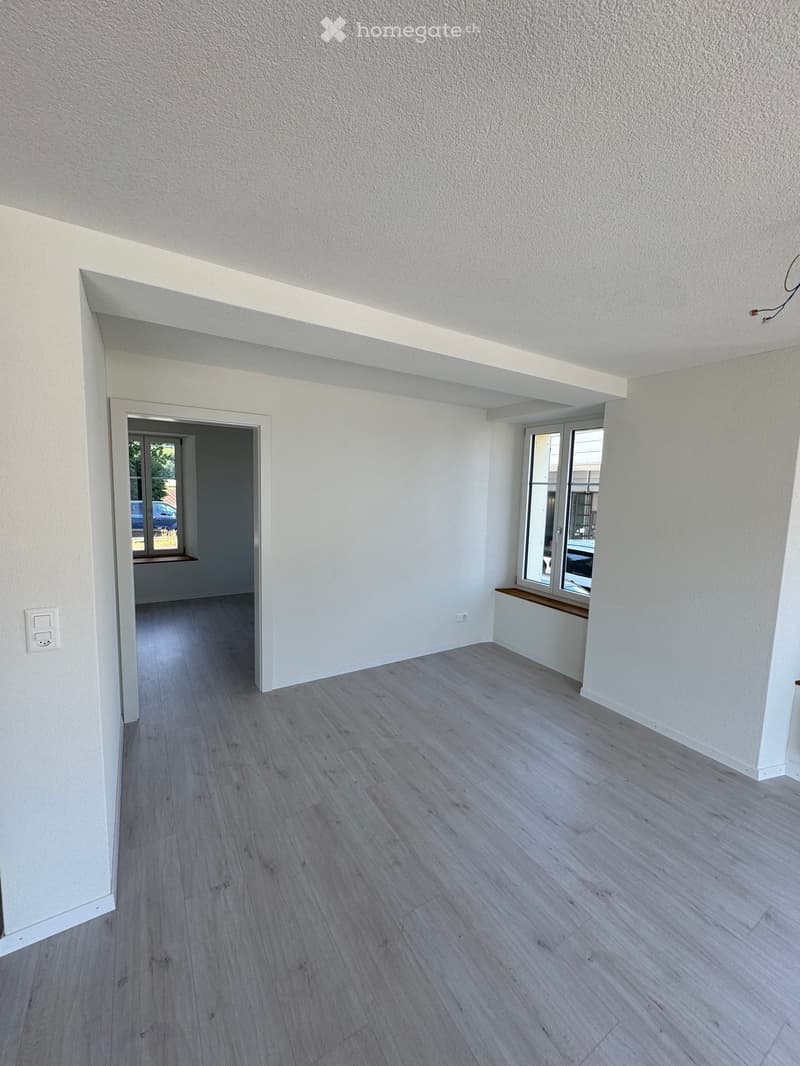 Empty room with light grey wooden flooring, white walls, and large windows allowing natural light. There is a doorway leading to another room with a window and a small bench.