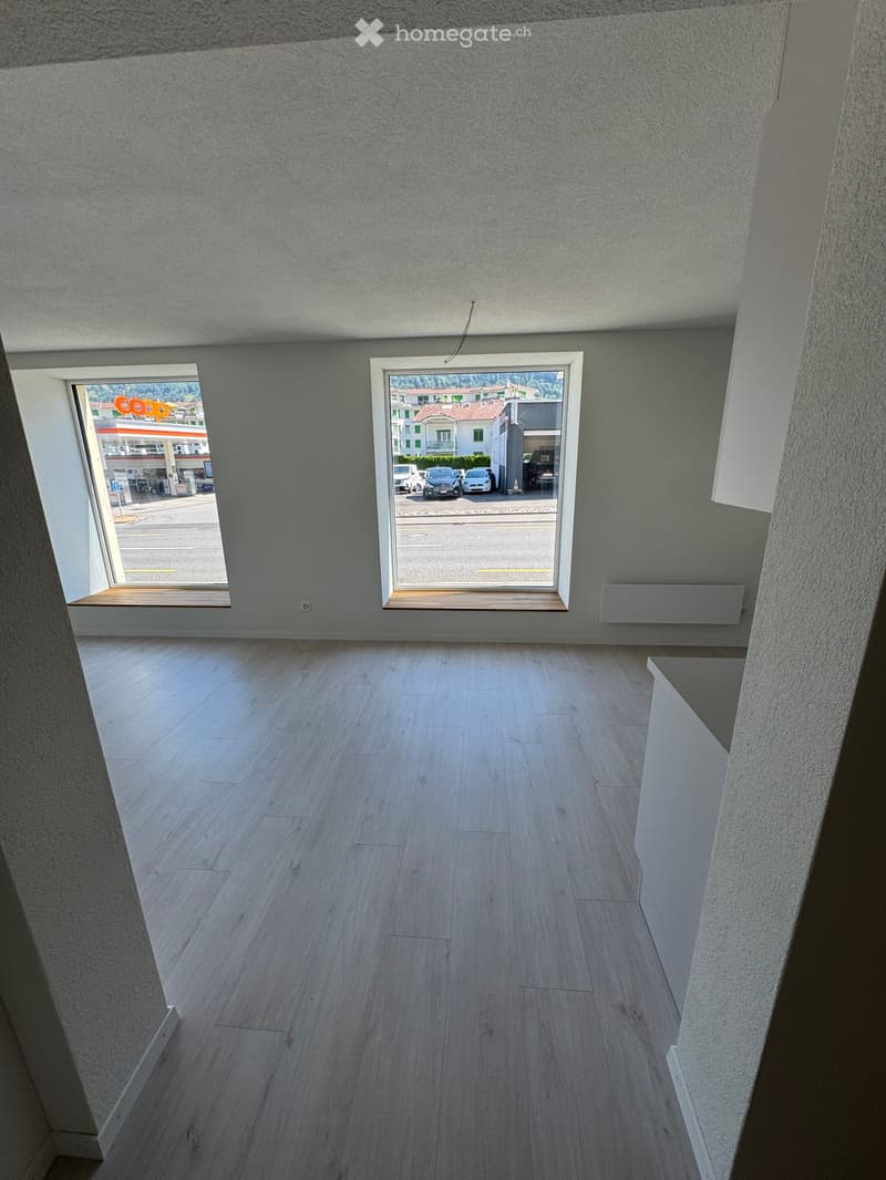 Empty room with light grey walls, wooden flooring, and large windows offering views of the street and nearby buildings.