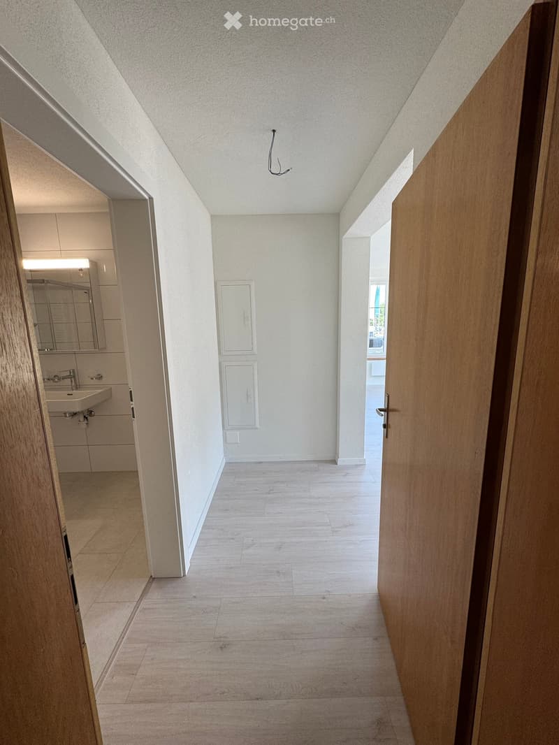 A narrow hallway with light-colored tiled flooring and white walls, featuring a partially open wooden door on the right and a closed door on the left leading to a small bathroom with a sink and mirror.