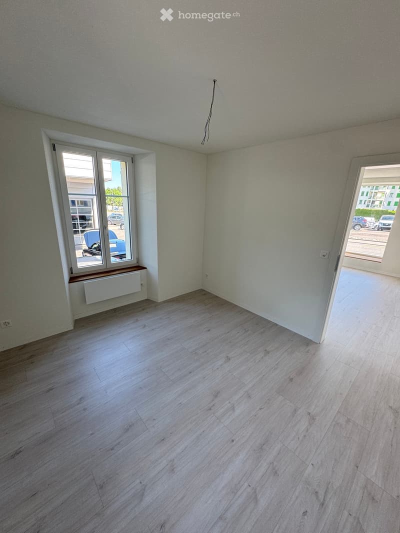Empty room with light-colored wooden flooring, white walls, and large windows offering a view of the street and parked cars.