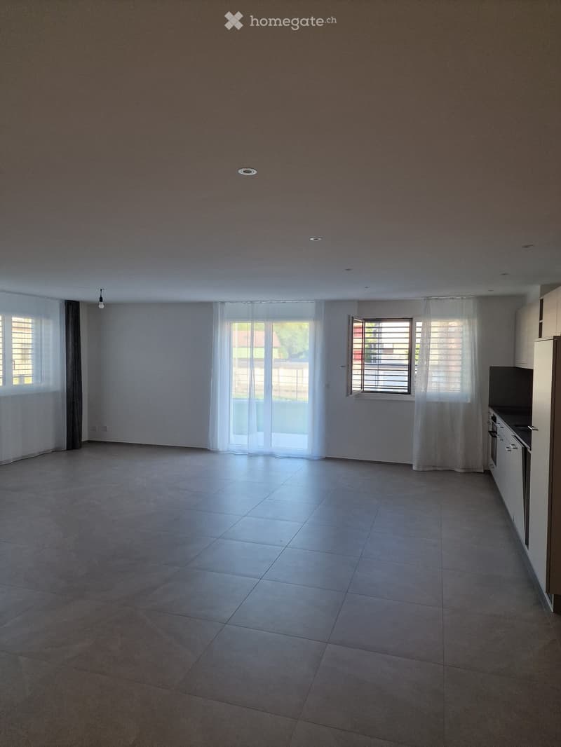 Empty room with tiled floor, white walls, large windows with white curtains, and a small kitchen area with cabinets and appliances visible on the right side.
