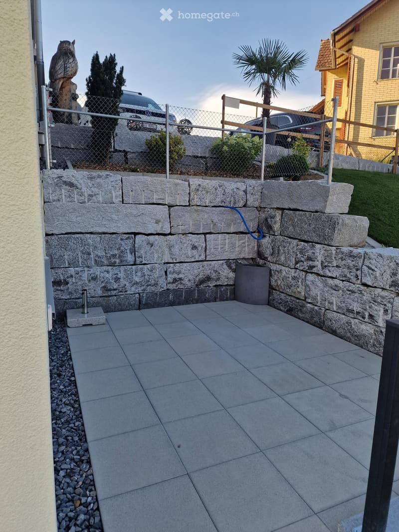 Raised stone platform with tiled surface, surrounded by gravel and a metal fence, with a palm tree and parked cars in the background.