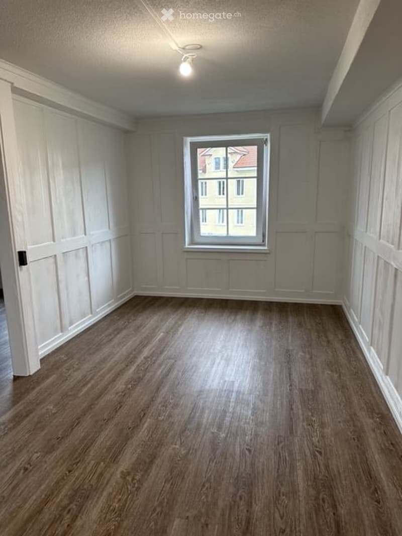 Empty room with wooden flooring, white paneled walls, and a single window showing a view of a neighboring building.