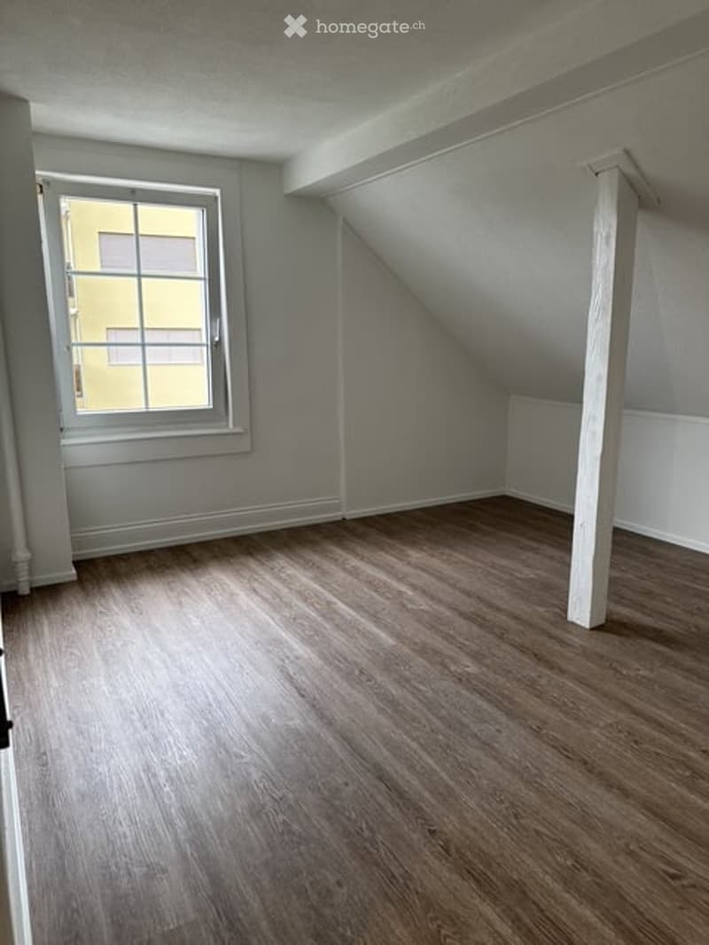 Empty attic room with wooden flooring, slanted ceiling, and a single window letting in natural light.