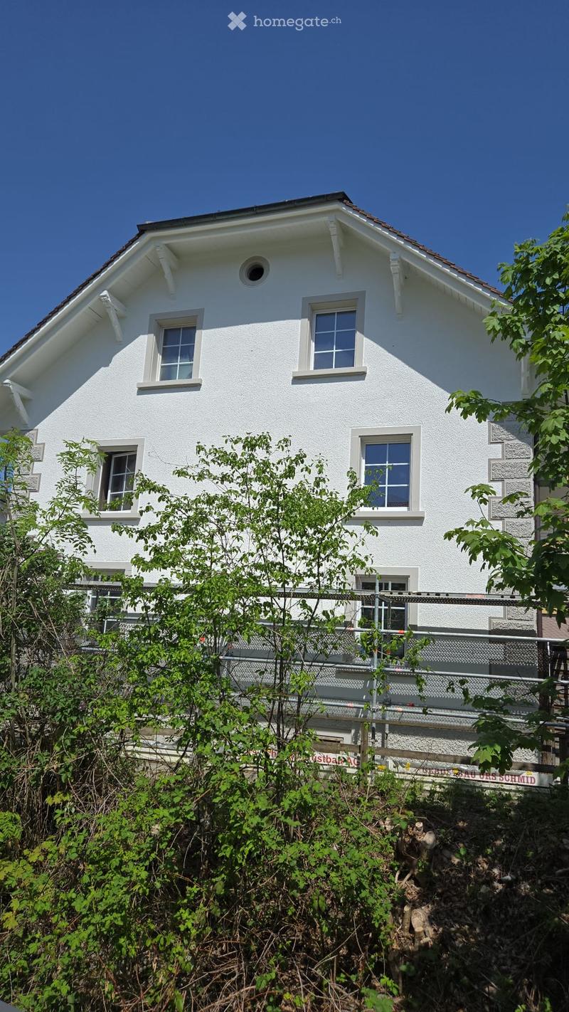 Two-story white house with multiple windows, surrounded by greenery and a fence.